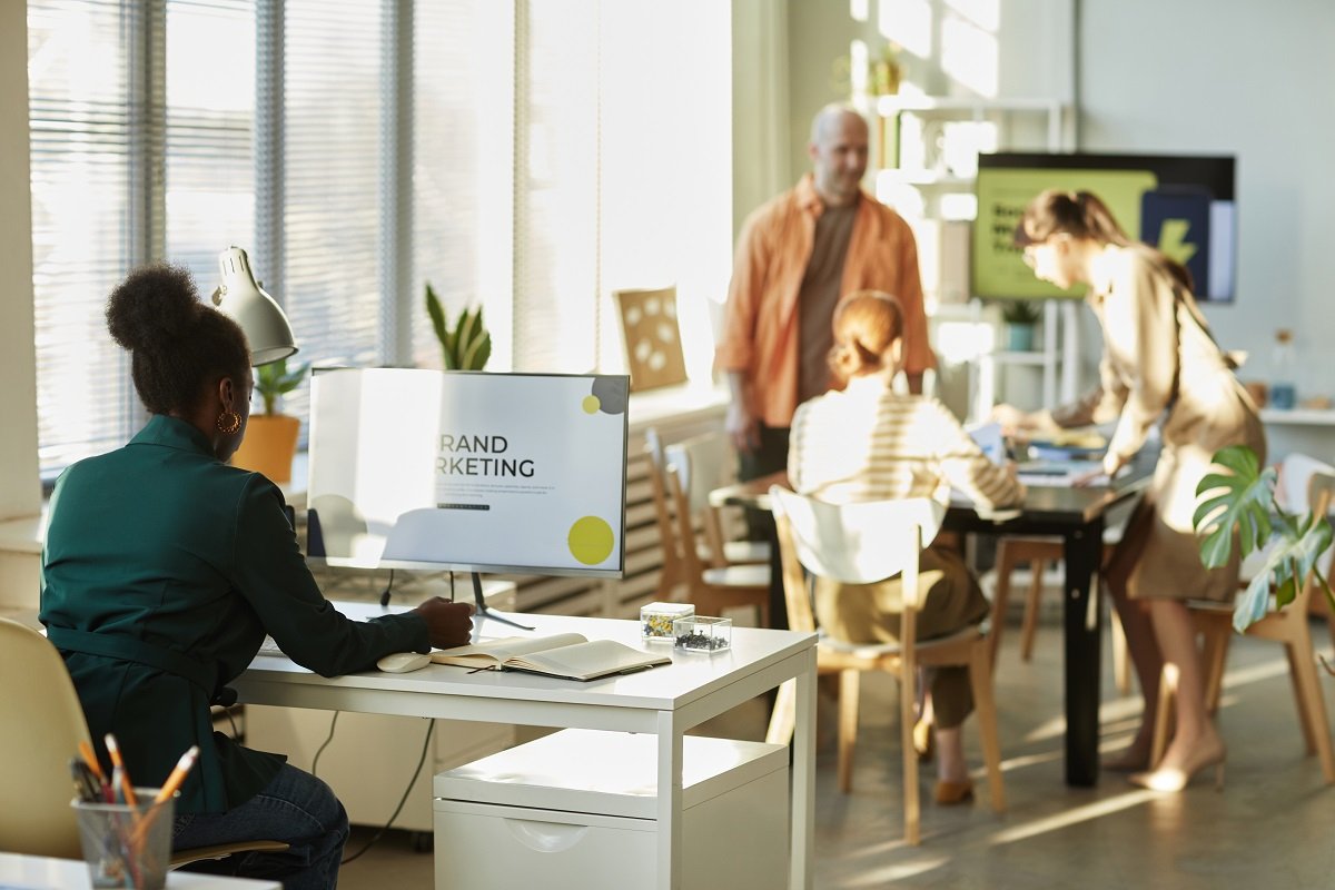 Three people sit around a table in an office while another works on brand marketing on a computer.