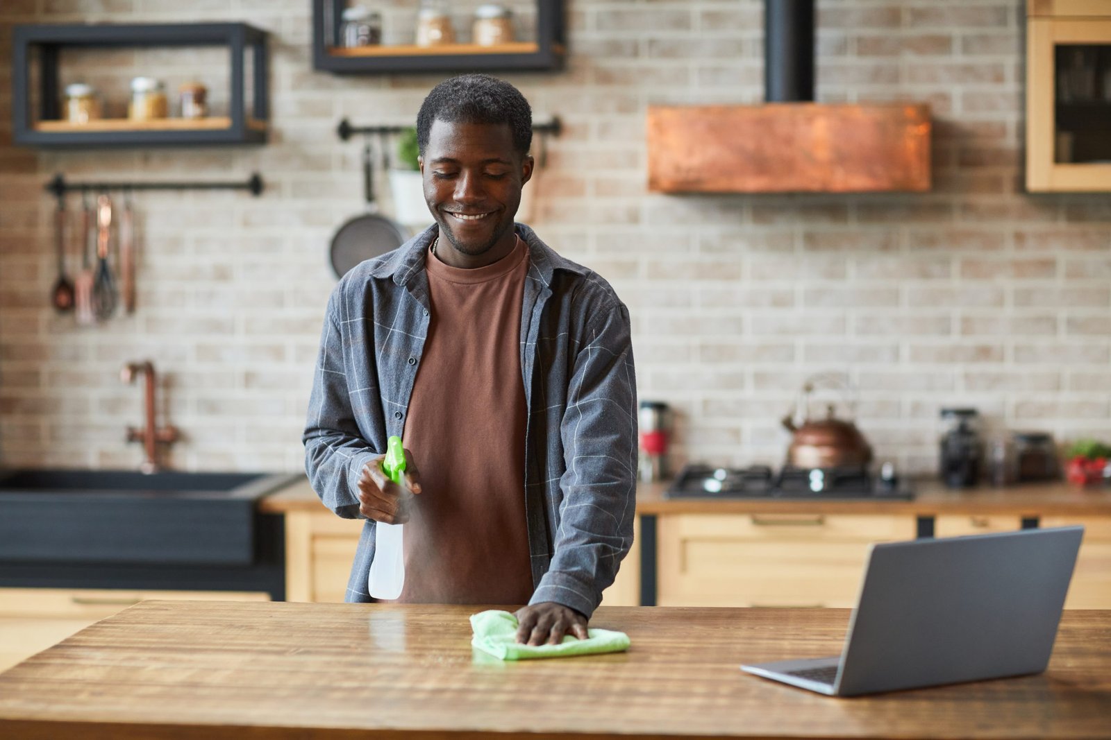 A man spring cleaning a kitchen counter with a laptop nearby.