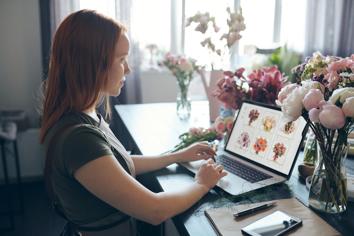 A woman sitting at a table full of vases of flowers looks at a laptop showing a website selling bouquets of flowers.